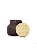 A reddish dark brown matte candle jar with a lit flame, shown next to its round gold-toned lid which features an embossed geometric floral pattern.
