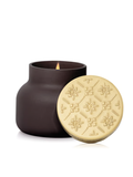 A dark brown matte jar candle with a lit flame, shown next to its round cream-colored lid which features an embossed floral pattern.