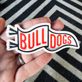 A hand holding a die-cut sticker that reads 'BULL DOGS' in red letters on a white banner with a black outline, set against a black and white striped rug.