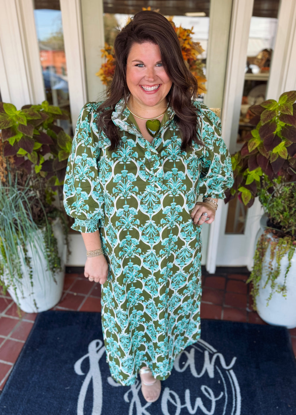 A woman stands smiling in front of a doorway wearing a long-sleeve midi dress with a green, teal, and white botanical pattern. She is standing on a dark blue doormat with white script lettering, flanked by potted plants.