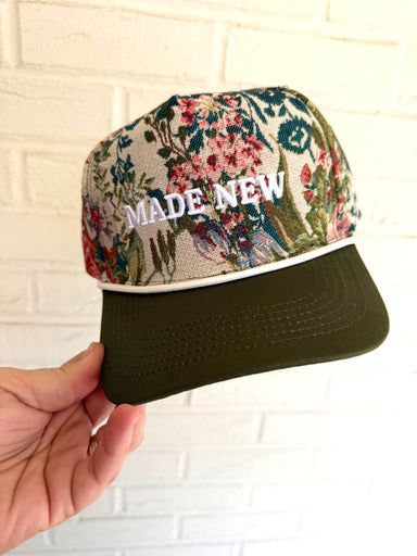 Floral-patterned cap with 'MADE NEW' text held against a white brick wall.