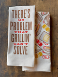 Two kitchen towels on a wooden surface, one with text and the other with grill-themed graphics.