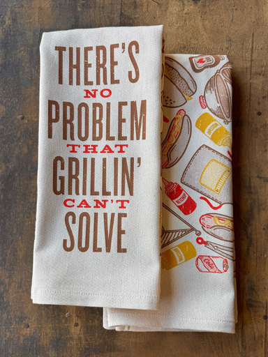 Two kitchen towels on a wooden surface, one with text and the other with grill-themed graphics.