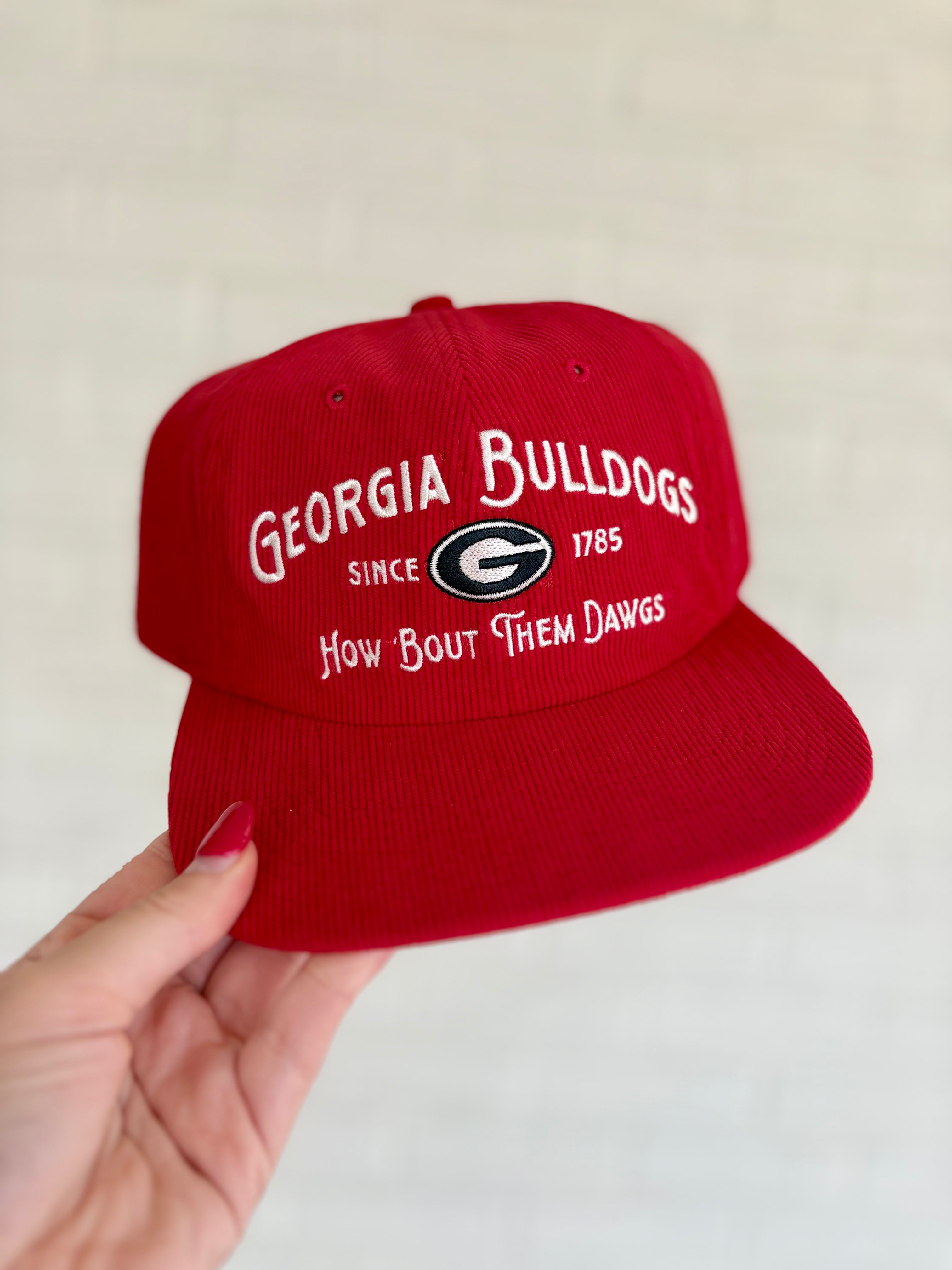 A red baseball cap with white embroidered text reading 'GEORGIA BULLDOGS', 'SINCE 1785', and 'How bout them dawgs' around a Georgia Bulldogs logo