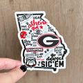 A hand with black nail polish holds a die-cut sticker shaped like the state of Georgia, featuring Georgia Bulldogs-themed text and graphics including the large 'G' logo, bulldog illustration, paw prints, and slogans, against a light wood background.