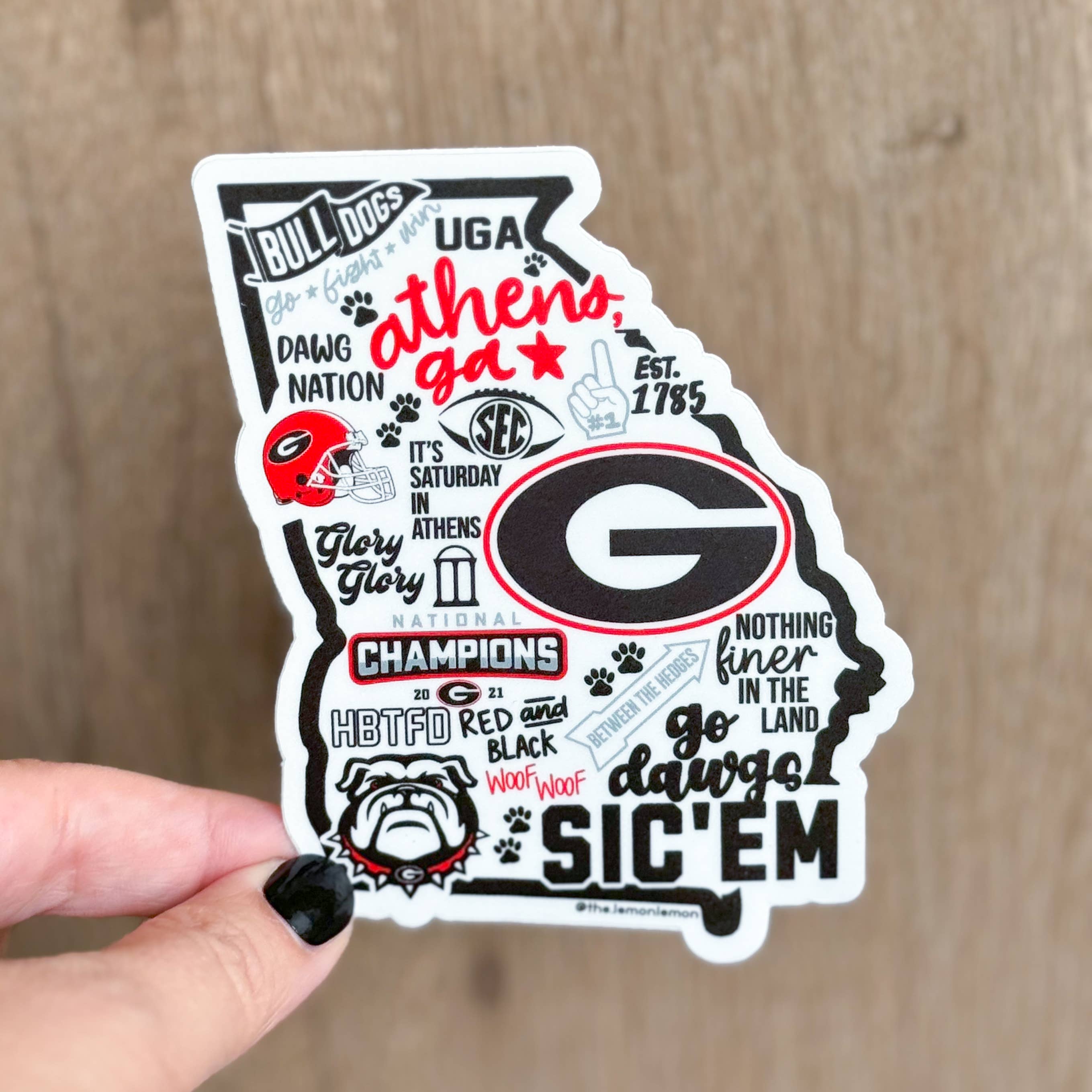 A hand with black nail polish holds a die-cut sticker shaped like the state of Georgia, featuring Georgia Bulldogs-themed text and graphics including the large 'G' logo, bulldog illustration, paw prints, and slogans, against a light wood background.