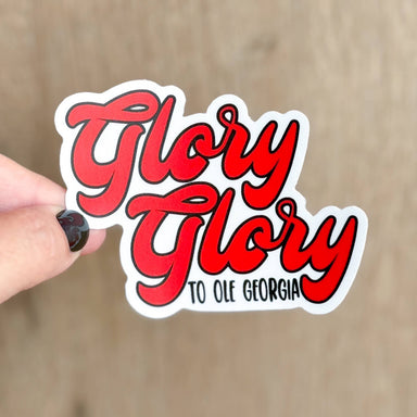 A hand with black nail polish holds a die-cut vinyl sticker that reads 'Glory Glory TO OLE GEORGIA' in red script with black outlines and a white border, against a blurred light brown wood background.