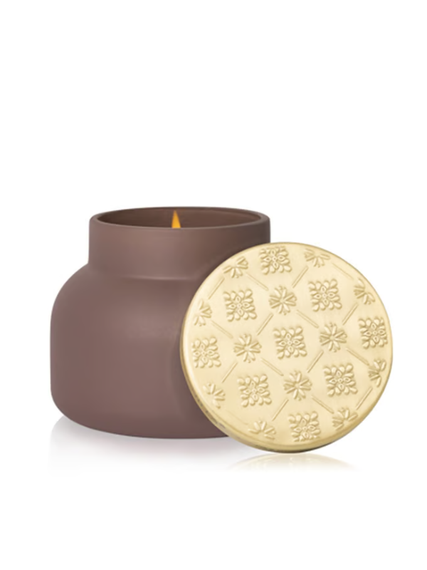 A brown matte jar candle with a lit wick, shown next to its round gold-toned lid which features an embossed floral pattern.
