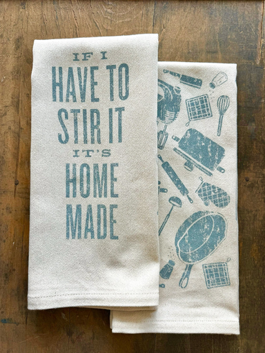 Two kitchen towels on a wooden surface, one with text and the other with printed designs.