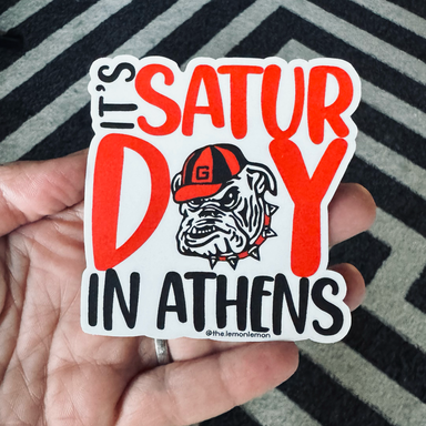 A hand holds a die-cut sticker that reads 'IT'S SATURDAY IN ATHENS' with a bulldog wearing a cap. The sticker is shown against a black and gray chevron-patterned rug.