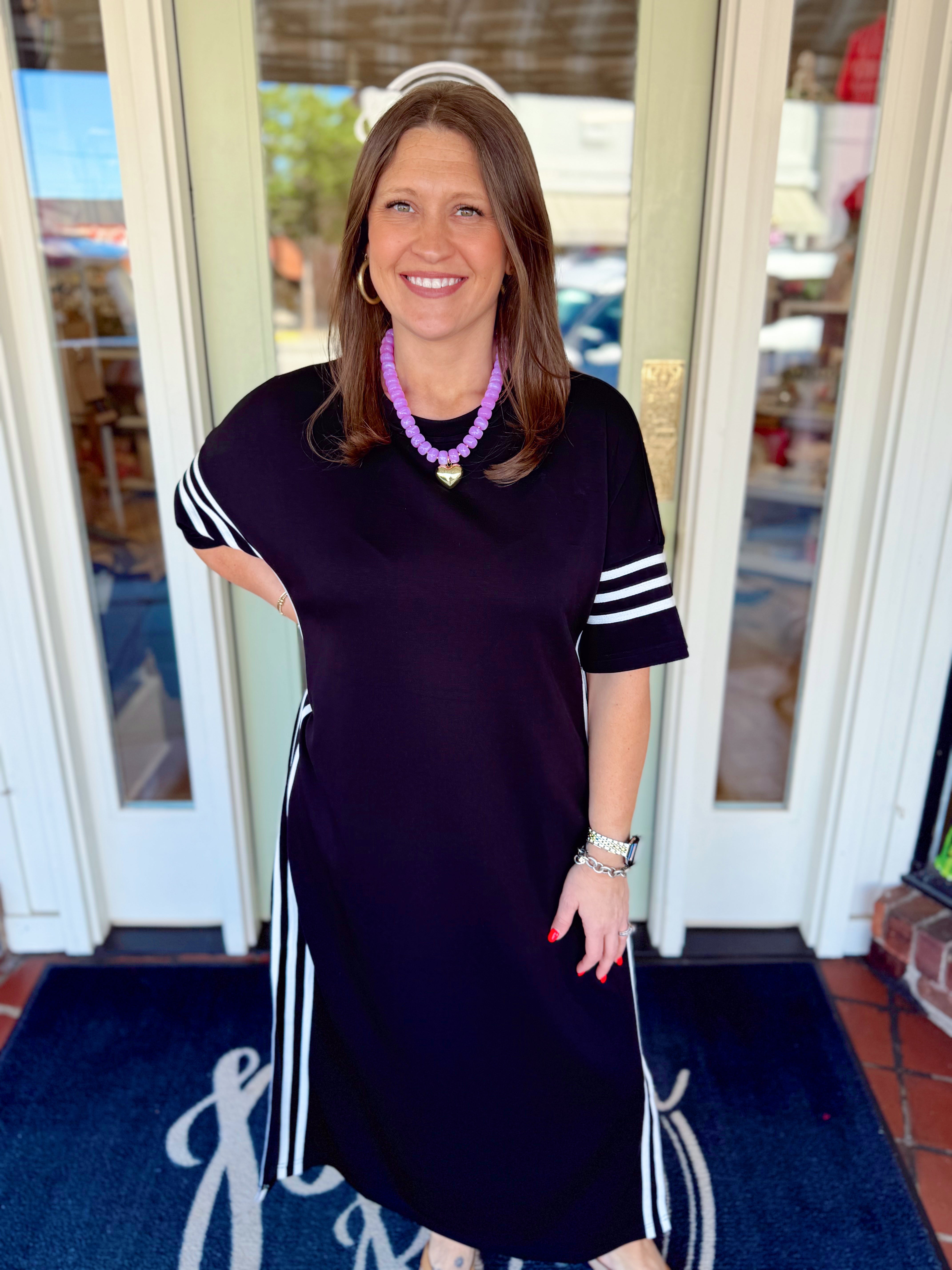 Woman wearing a black midi dress with white striped sleeve and side details paired with a purple beaded necklace and sandals.
