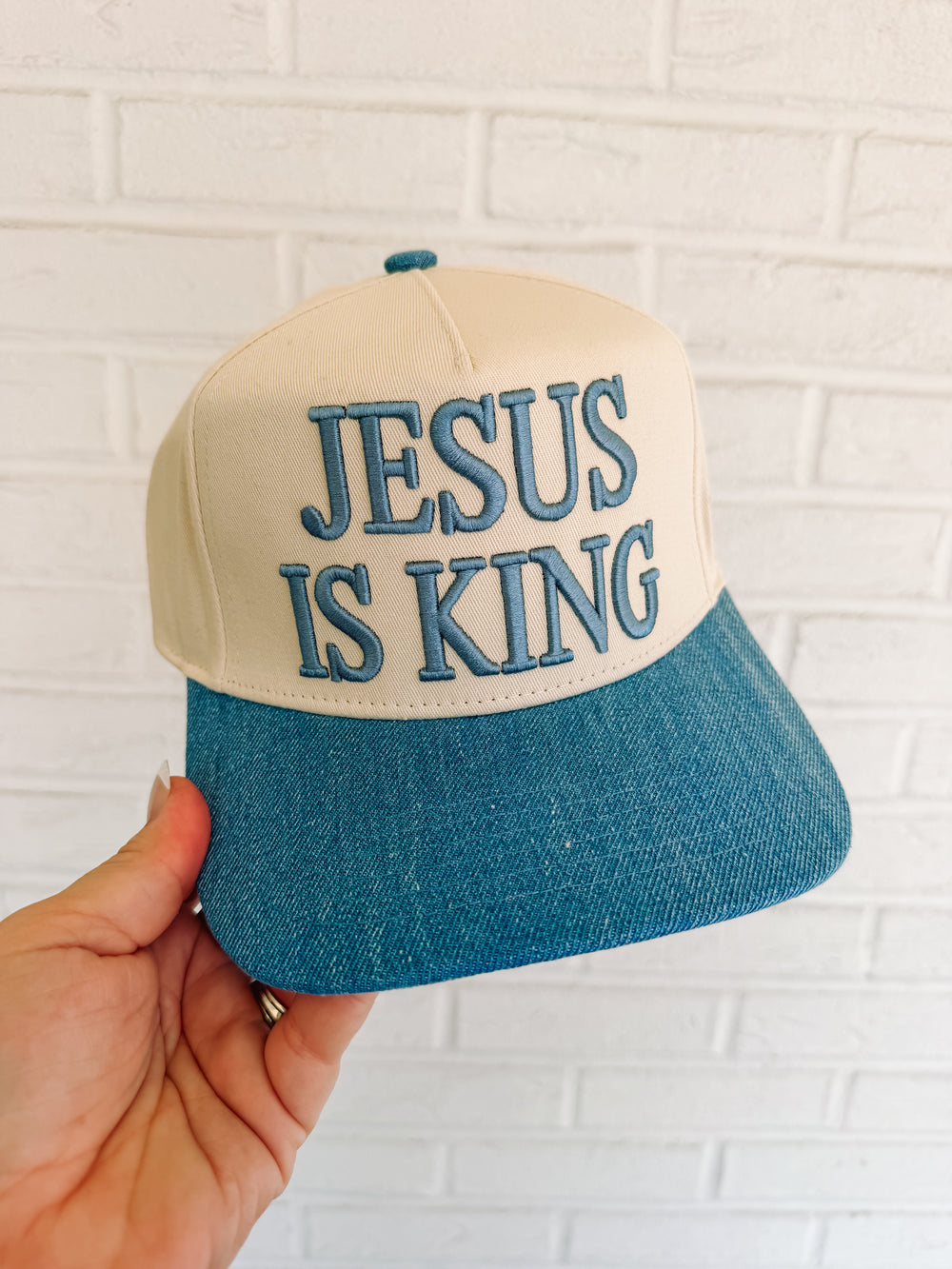 Cap with 'Jesus is King' text held against a white brick wall.