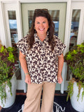 A smiling woman stands in front of a glass door wearing a short-sleeve leopard-print button-up shirt and tan pants, with potted plants on either side.