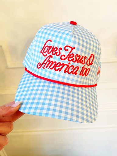 Blue and white checkered cap with red text 'Loves Jesus & America too' held by a hand against a light background
