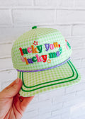 Green checkered cap with colorful text held against a white brick wall.