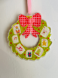 A festive Christmas wreath-shaped ornament hanging on a white wall. The ornament features a bright green scalloped border with red polka dots, a large red and white gingham bow at the top, and six numbered advent cards (1 through 6) attached around the wreath.