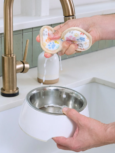 A person holds a white pet water bowl with a stainless steel interior under a brass faucet, while another person squeezes a floral-patterned pet lick mat above the bowl to wet it in a sink setting.