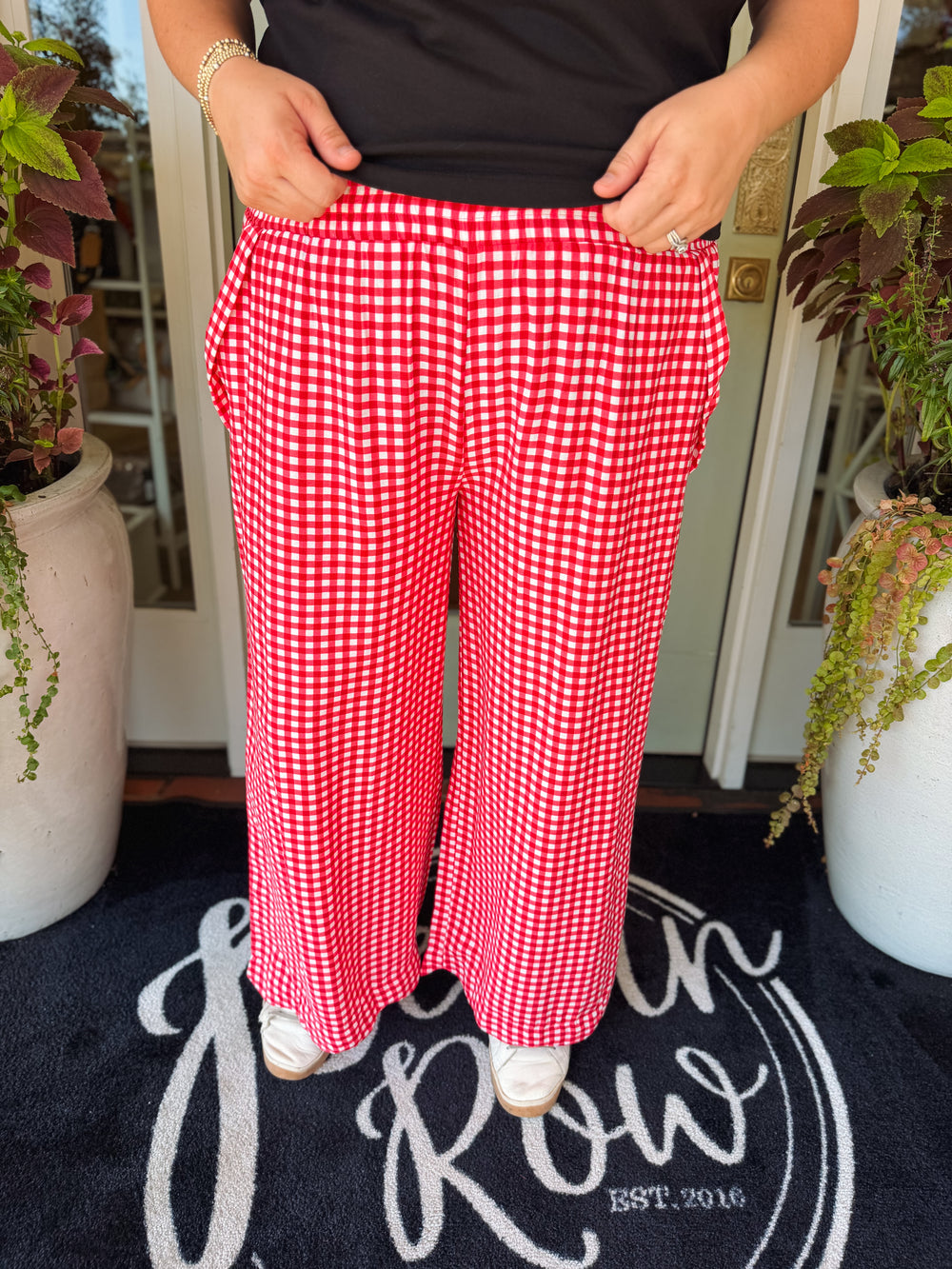 A person wearing red and white gingham wide-leg pants, standing on a black doormat in front of a white door with potted plants on both sides.