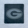 A square black slate coaster featuring a debossed gray Georgia Bulldogs 'G' logo, set against a light gray textured fabric background.