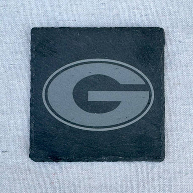 A square black slate coaster featuring a debossed gray Georgia Bulldogs 'G' logo, set against a light gray textured fabric background.