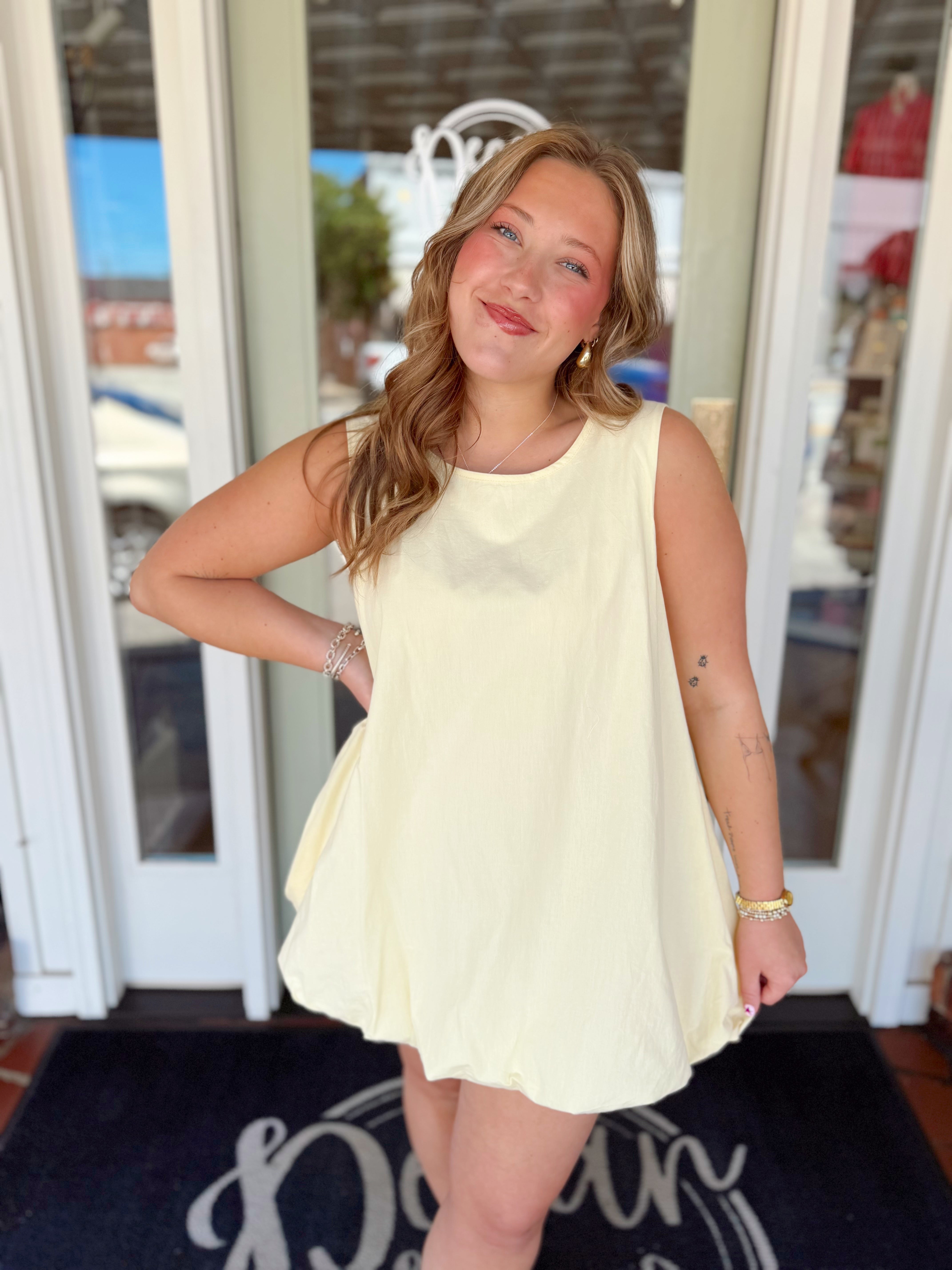 Woman wearing a sleeveless soft yellow mini dress with a relaxed swing silhouette and bubble hem detail.
