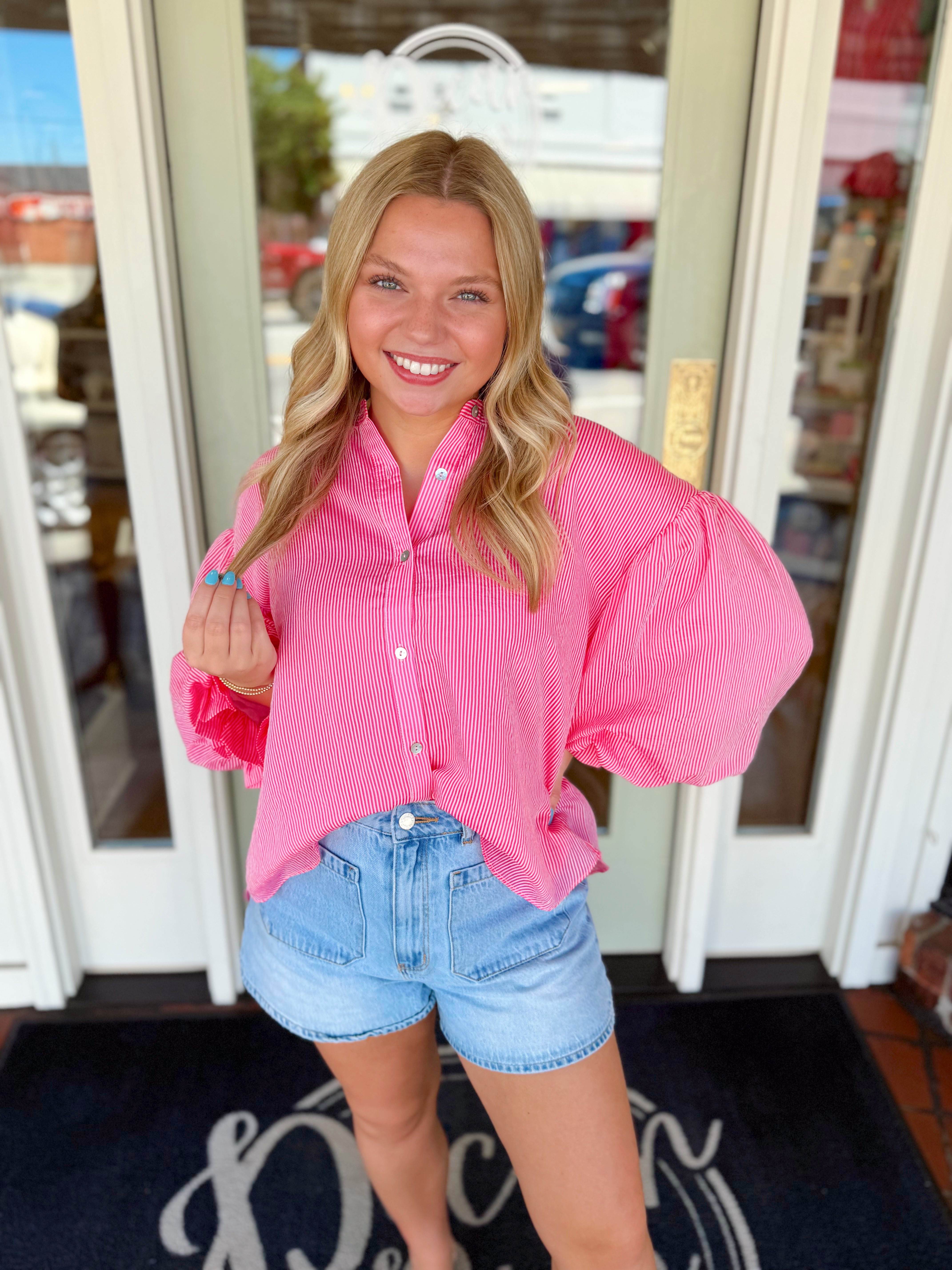 Woman wearing a pink striped button-down blouse with statement puff sleeves paired with light wash denim shorts.
