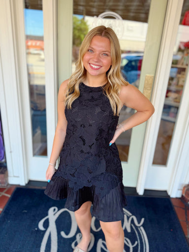 Woman wearing a sleeveless black floral lace mini dress with a ruffle hem and relaxed silhouette.

