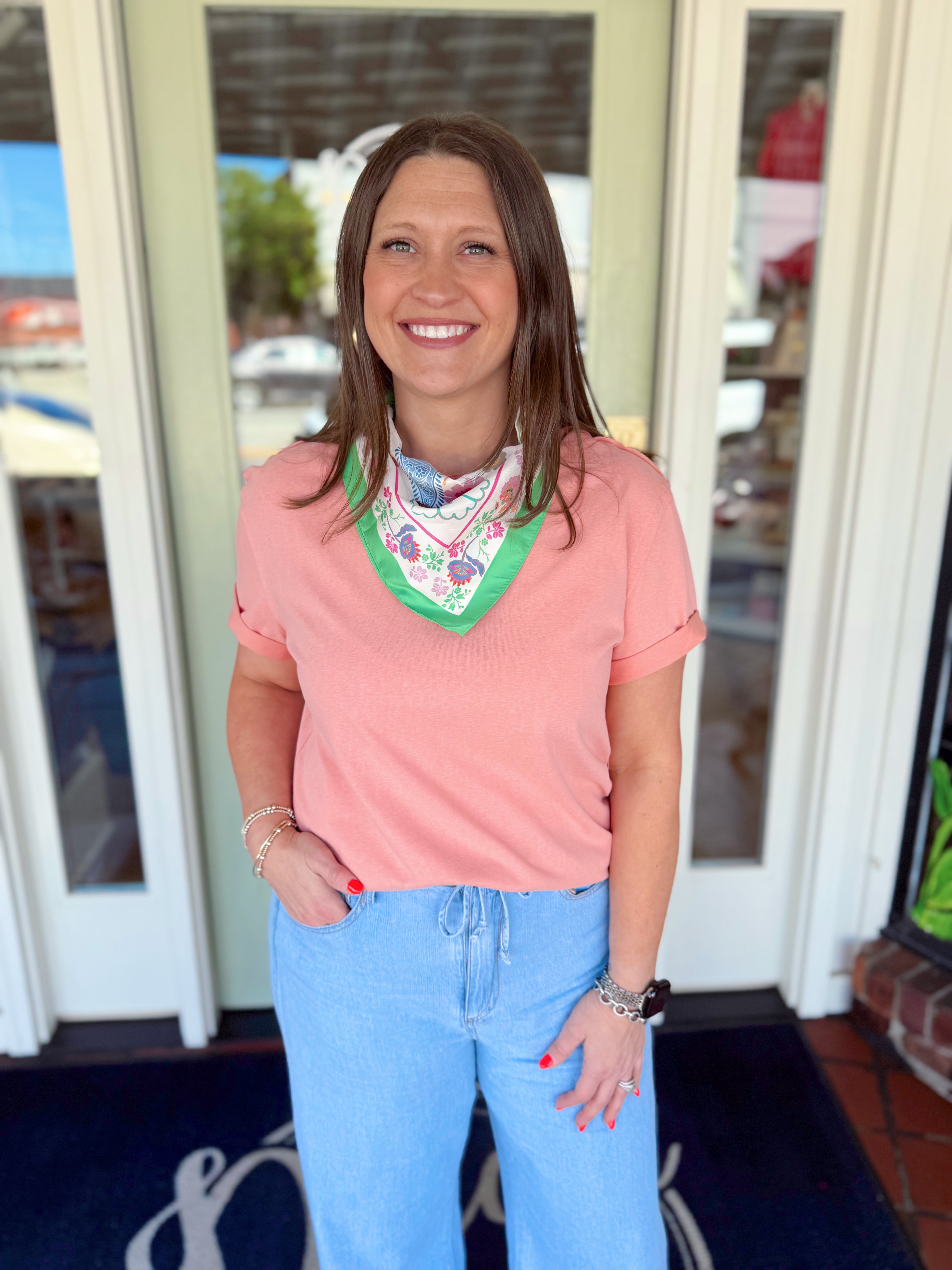 Woman wearing a blush pink short sleeve tee with rolled sleeves paired with light wash denim jeans and a colorful floral neck scarf.