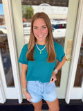 Woman wearing a deep teal short sleeve tee paired with light wash denim shorts and a white beaded necklace with green pendant detail.
