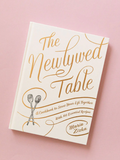 A cookbook titled 'The Newlywed Table' by Maria Zizka, featuring a white cover with gold lettering and illustrations of crossed spoons.