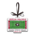 A Georgia Bulldogs ornament shaped like a football field, with a dark brown ribbon loop at the top. The design features a green field, white yard lines, red side banners reading 'GEORGIA' and 'BULLDOGS', a large 'G' logo in the center, and text reading 'GEORGIA BULLDOGS' and 'MAKING SPIRITS BRIGHT SINCE 1784'.