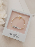 Bracelet with 'WORTHY' charm in a box with 'I am WORTHY' text, surrounded by flowers.