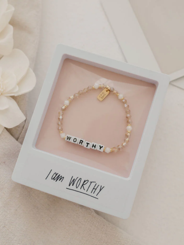 Bracelet with 'WORTHY' charm in a box with 'I am WORTHY' text, surrounded by flowers.