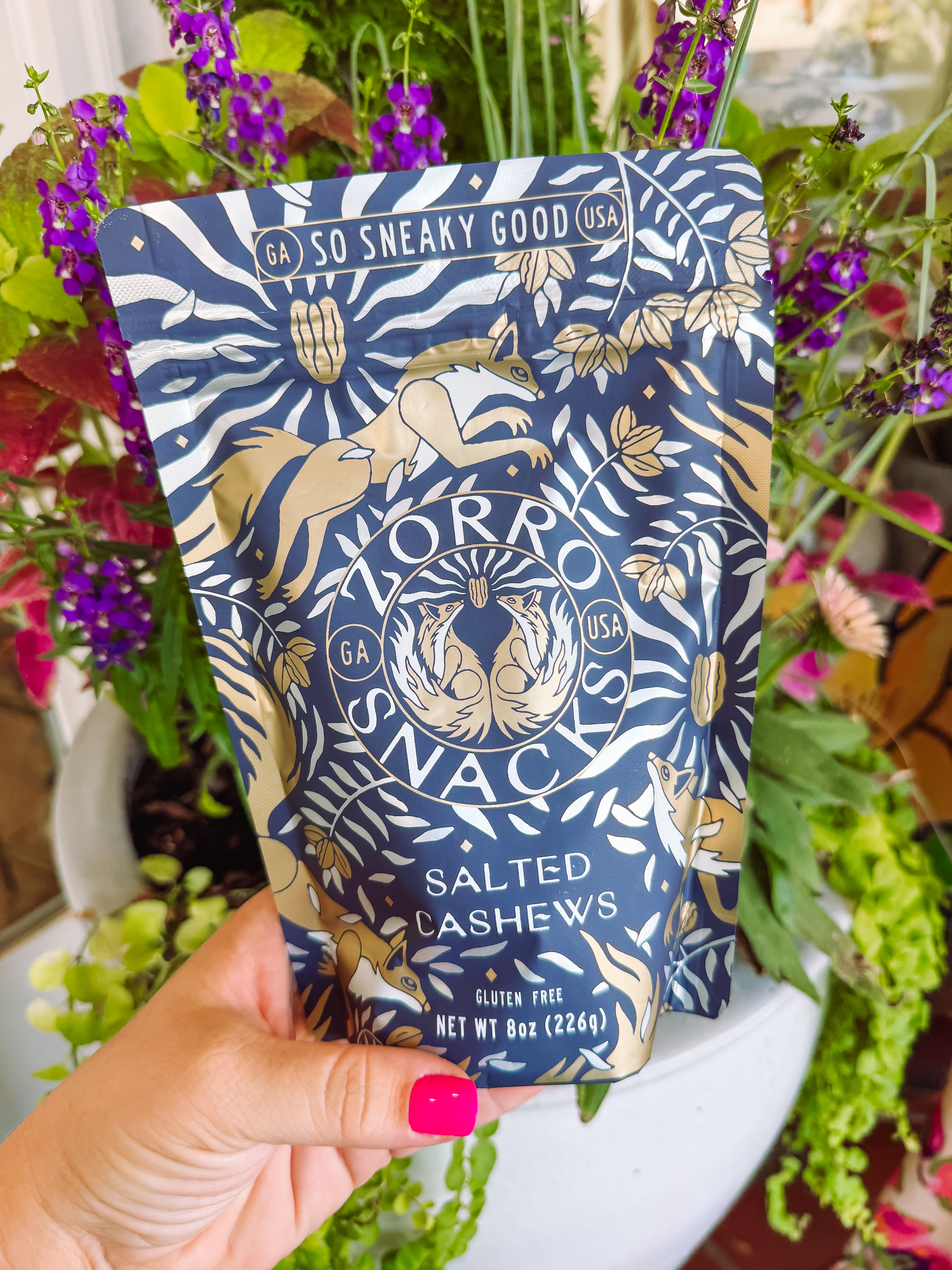 A hand with bright pink nail polish holds a stand-up pouch of Morro Snack Salted Cashews. The pouch features a navy blue background with a gold foil pattern of a tiger and zebra stripes.