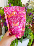 A hand holds a bright pink and orange stand-up pouch of Zorro Pecano Sweet & Spicy coffee against a background of green potted plants.