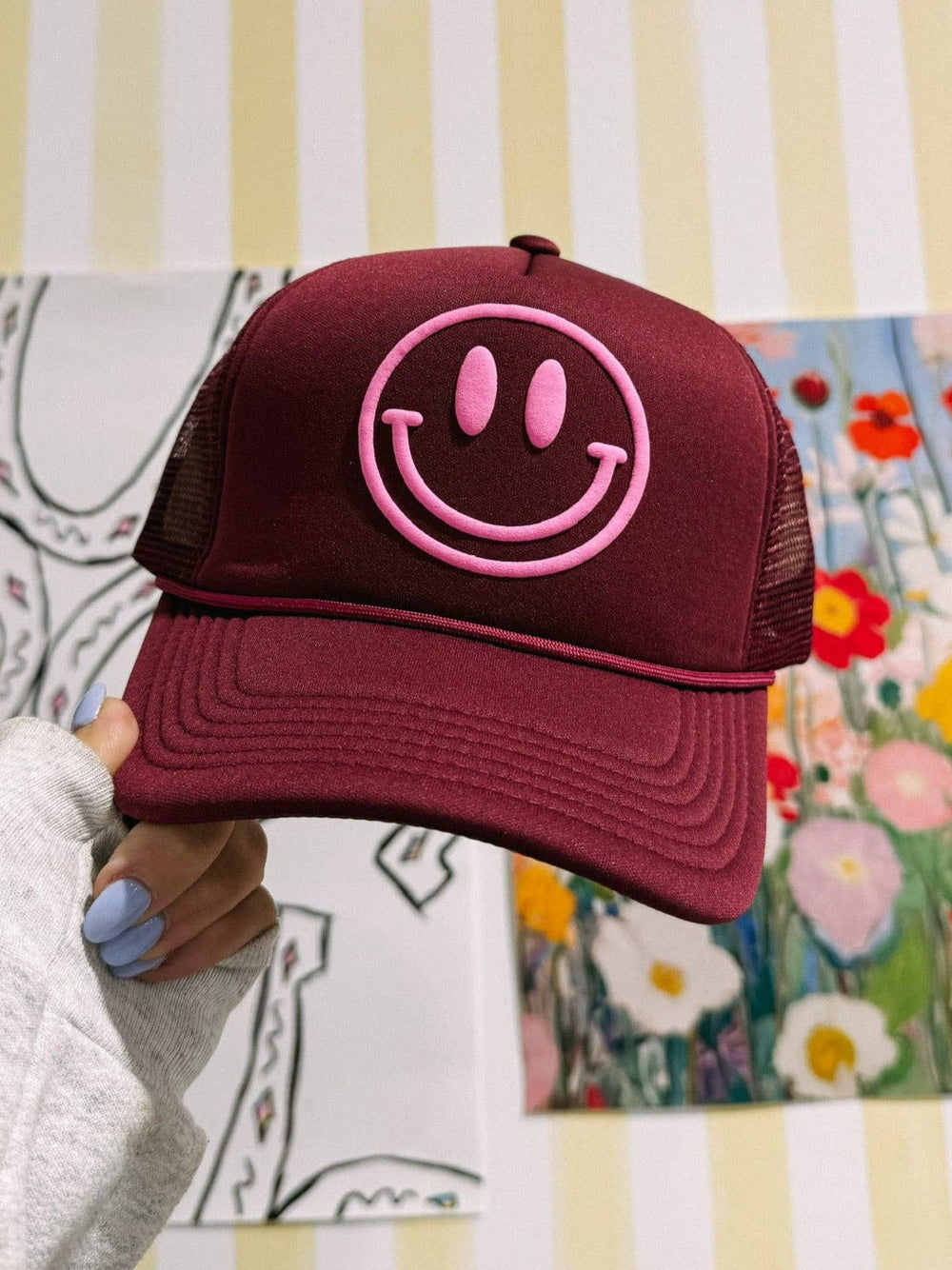 Maroon cap with pink smiley face held against a colorful and patterned background