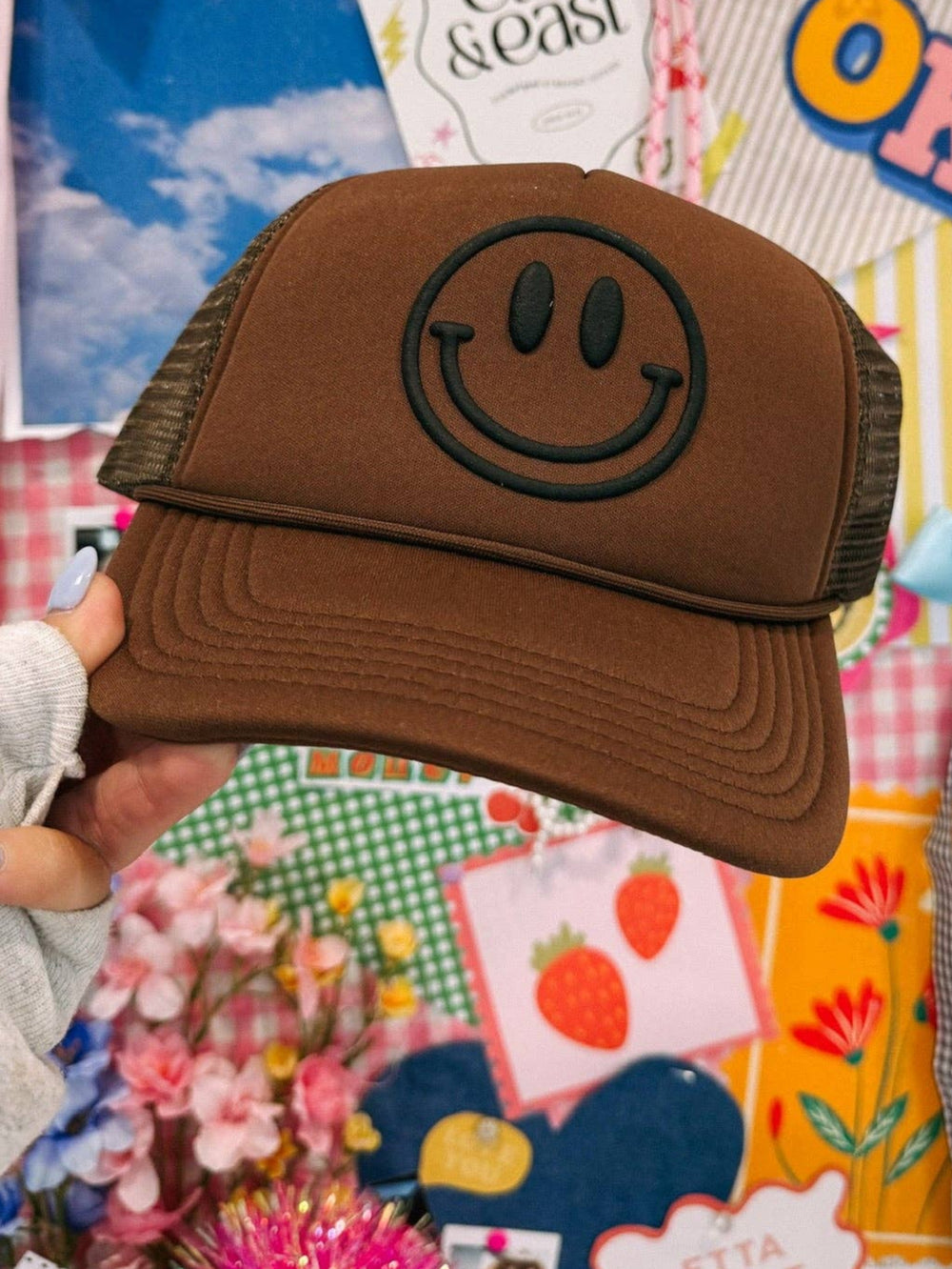 Brown cap with smiley face design held in front of a colorful collage background