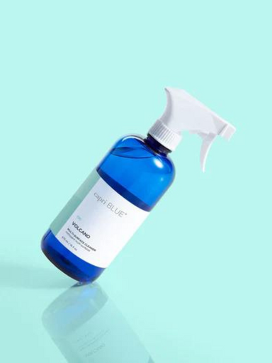 A blue spray bottle of Igari BLUE Volcano facial cleanser with a white trigger sprayer, set against a solid mint green background with a soft reflection below.